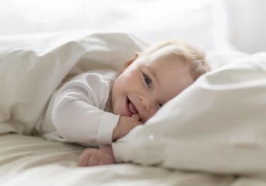 A happy, smiling newborn baby boy lying in bed under a white duvet cover.