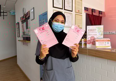 A healthcare worker wearing a mask holds pink maternal health records at a Malaysian medical clinic.