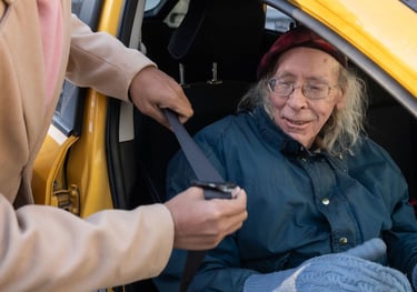 A caregiver helps an elderly person fasten their seatbelt in a yellow car for senior transportation services.