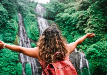 A female traveler with a red backpack stands with arms wide open before a lush tropical waterfall.