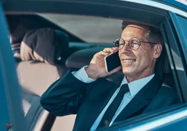 Smiling businessman in a suit talking on a smartphone while riding in the back of a luxury car.