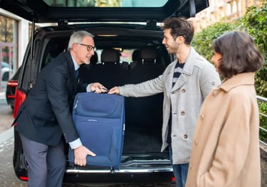 Professional chauffeur assisting a young couple by loading blue luggage into a black van.