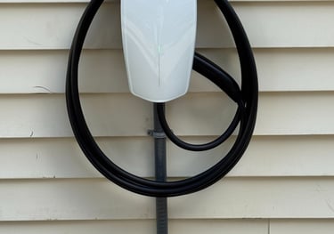 White Tesla Wall Connector home charging station mounted on beige vinyl house siding with coiled black cable.