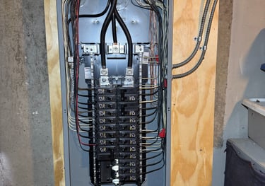 Open residential electrical panel with circuit breakers and wired connections mounted on a plywood wall.
