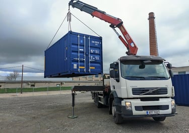 A Volvo crane truck lifting a blue shipping container at an industrial logistics site.