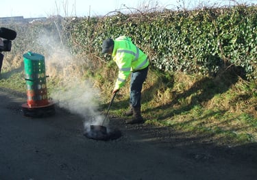 A worker in high-visibility gear repairs a pothole on a rural road using steaming asphalt.