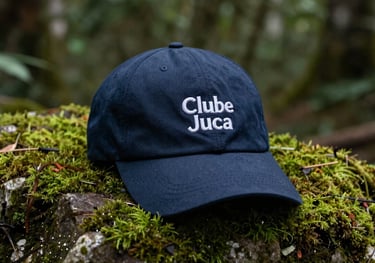 A dark blue cap with the Clube Juca logo embroidered on the front, placed on a mossy rock in a South American / Brazilian forest setting.