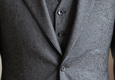 A close-up photograph of a three-piece suit in soft gray wool, showcasing the precision of the stitching and the luxurious texture of the fabric, International / Western.