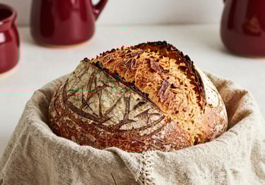 A high-quality photo of a basket of freshly baked artisanal sourdough bread resting on a rustic cloth. The setting is a bright, sunlit Brazilian kitchen with hints of Deep Carmine Red in the ceramics.