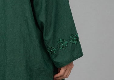 A close-up photography of a forest green linen abaya sleeve featuring delicate hand-stitched floral embroidery. The fabric has a natural, premium texture. Soft-focus background with English color name warm grey. Middle Eastern / Gulf style.