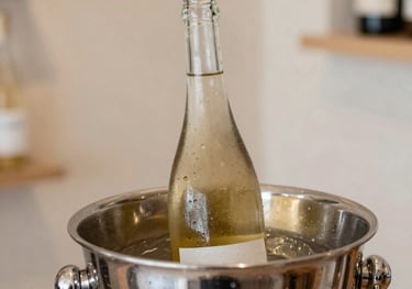 A close-up of a chilled white wine bottle in a modern silver ice bucket. Condensation beads on the glass. The setting is a bright, airy boutique shop corner with light sand-colored walls.