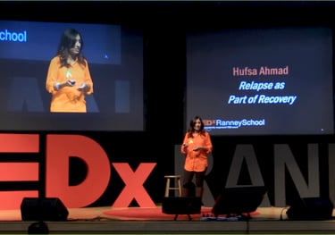 A woman in an orange shirt speaks energetically on stage. The screen reads "Hufsa Ahmad: Relapse as Part of Recovery."