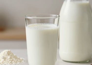 A close-up of a glass of rich, creamy milk standing next to a container of powder milk. The lighting is soft and natural, emphasizing purity and health in an off-white domestic kitchen setting.
