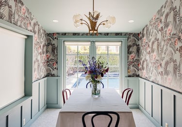 Modern dining room featuring tropical floral wallpaper, sage green wainscoting, and a view of a backyard pool.