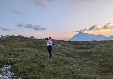 Laura stands on a green field at sunset taking a photo. The Italian Dolomites are in the background