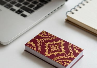 A high-angle shot of a neat office desk with a laptop, a notebook, and a business card featuring deep crimson and gold batik motifs.