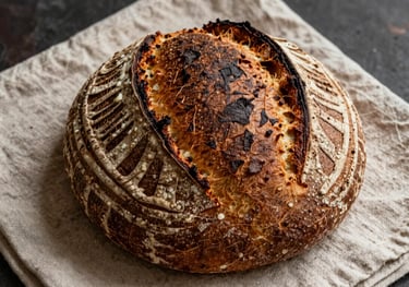 A high-angle photo of a large sourdough loaf on a linen napkin, showing deep scoring marks and a dark brown, crunchy exterior. Rustic and artisanal vibe.