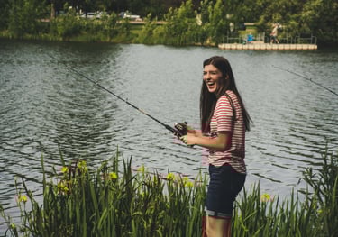 mulher morena com blusa listada pescando com vara de fibra e lago ao fundo.