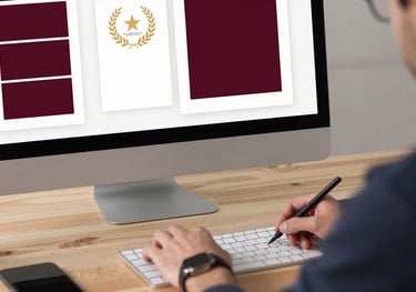 A professional designer at a clean wooden desk, working on a book cover layout on a large monitor. A mood board with deep ripe crimson swatches is visible.