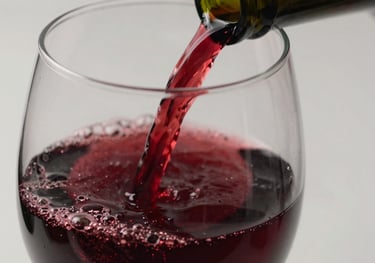 Close-up of a glass of dark red wine being poured, capturing the rich texture and deep crimson color, against a neutral white background.