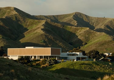 A serene and professional photo of a New Zealand landscape, featuring rolling green hills and modern architecture in warm tan and soft off-white.