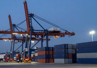 An evening shot of a busy harbor terminal in North America with large cranes and stacked blue and gray shipping containers under bright stadium lights.
