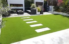 Modern garden with lush artificial grass, white stepping stones, and black outdoor lounge furniture.