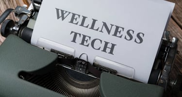 A typewriter with a sheet of paper in the carriage displaying the text 'WELLNESS TECH'. The typewrit