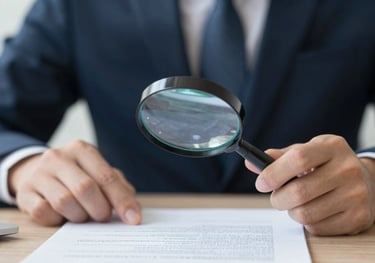 A person in professional attire reviewing an official document with a magnifying glass, suggesting detail-oriented validation. Clean, high-end look.