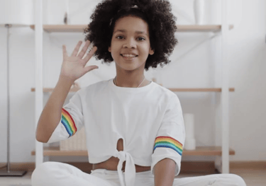 Little girl in a white pant set, sitting facing the camera waving hello