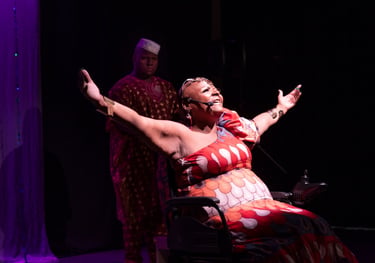 Fotografia de cena teatral com a artista Mona Rikumbi. Mulher negra, usuária de cadeira de rodas 