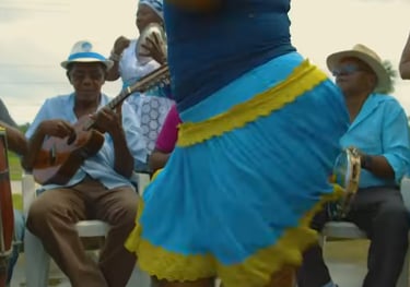 Grupo de samba de roda ao ar livre, com uma sambadeira vestida de azul ao centro e músicos ao redor.