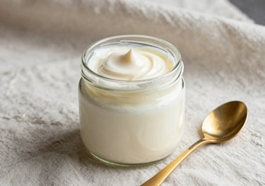 A glass jar of thick, clotted cream sitting next to a gold spoon on a light cream linen cloth, elegant and minimalist composition, focus on texture and purity.