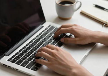 Detail of hands typing on a sleek, silver laptop keyboard. Beside the laptop is a small ceramic cup of coffee and a notebook. Minimalist desk setup with soft natural light in a professional study.
