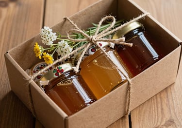 A beautifully presented gift box containing three jars of honey on a wooden surface, tied with natural twine and a wildflower sprig. Southeastern European / Albanian aesthetic.