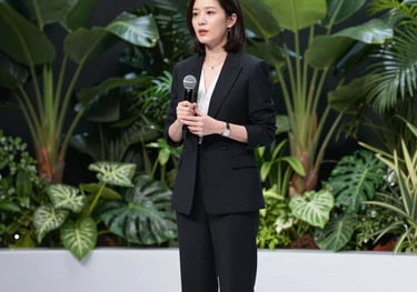 A professional woman presenting on a bright, minimalist stage with a lush botanical background. The lighting is soft and flattering, emphasizing a premium educational environment.