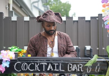 A male bartender in a pirate costume works at an outdoor tiki cocktail bar for a themed party.
