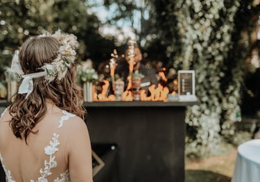 A bride with a floral crown watches a flair bartender perform a fire show at an outdoor wedding reception.