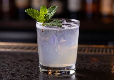 A close-up of a sophisticated cocktail garnished with fresh mint, sitting on a dark polished bar. The liquid reflects soft lavender gray and deep midnight indigo lights.