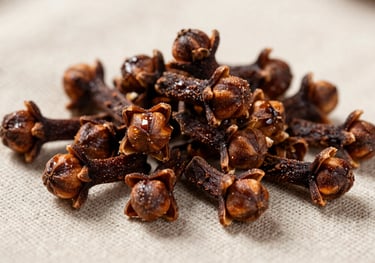 A pile of high-quality whole cloves on a neutral linen background. The photo is sharp, showing the rich dark brown color and oily sheen of the spice. Natural side lighting highlights the intricate shape of the flower buds.