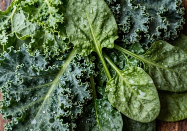 A top-down, high-quality photography shot of vibrant, mixed leafy greens like kale and spinach, misted with water for freshness. The composition is clean and organic, set against a rustic wooden background in a North American market style.