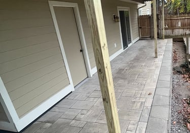 Custom stone paver patio installed under a raised wooden deck with light brown vinyl siding.