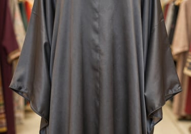Detailed photography of a premium silk abaya draped elegantly. The fabric color is a soft charcoal. Southeast Asian boutique setting with warm, soft-focus lighting.