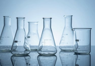 A collection of crystal-clear borosilicate laboratory flasks and beakers arranged on a reflective surface, reflecting medium blue and light blue light in a clean studio setting.