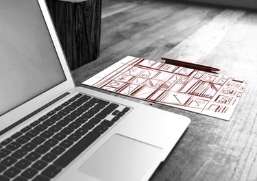 A laptop on a wooden desk next to a sheet of paper with red wireframe designs