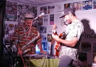 Dos músicos interpretan música en vivo con un saxofón y una guitarra acústica frente a una pared cub