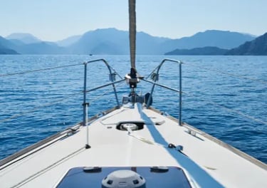 View from the bow of a white sailboat cruising on blue ocean water toward distant mountains.
