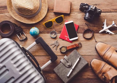 Travel essentials flat lay on a wooden table including suitcase, passport, camera, and hat.