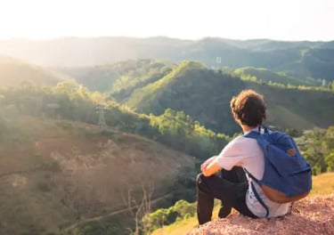 A male hiker with a blue backpack sits on a hill at sunset, looking over a lush mountain valley.