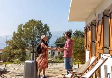 A smiling female traveler with a suitcase greeting a host outside a Mediterranean vacation rental.
